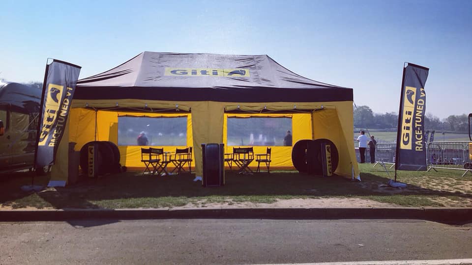 Rider sheltering with bike and kit under a race awning in the rain