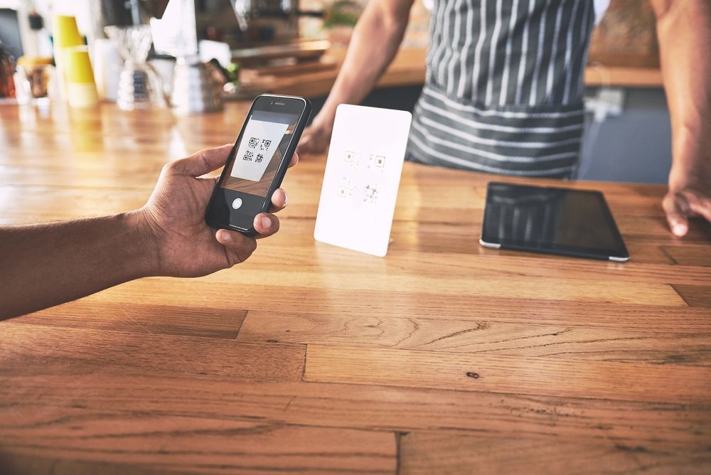Customer scanning a QR code on a countertop stand with their phone to pay, staff member behind the counter